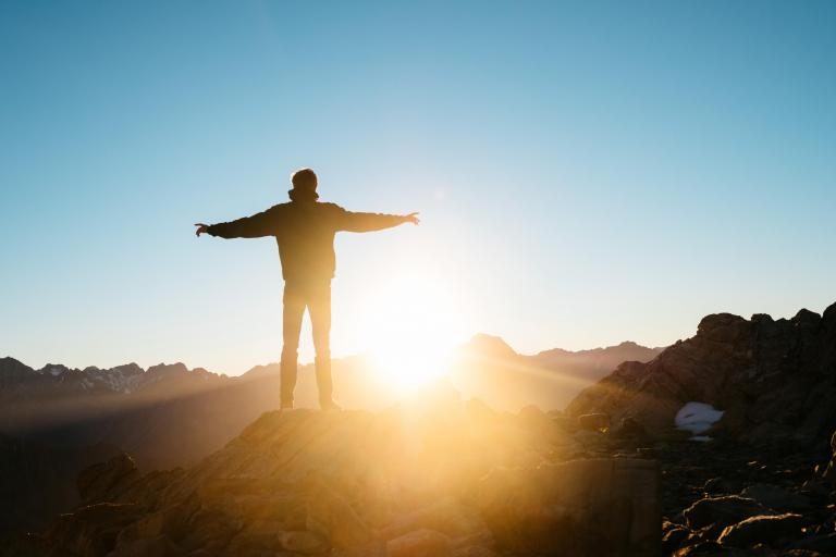 Foto van een man op een bergtop die zijn armen strekt tijdens zonsopgang